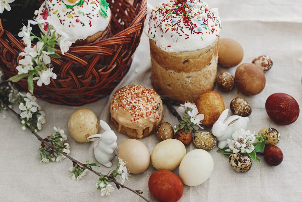Huevos de Pascua modernos, pan de Pascua, cesta de mimbre, conejito y flores de primavera en flor en mesa rústica. ¡Felices Pascuas! Tarta casera y huevos teñidos al natural. Comida tradicional