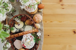 Delicious Easter food, stylish easter eggs, beets, cheese, butter, ham, homemade Easter bread in wicker basket with blooming spring flowers on rustic table, top view. Happy Easter! Orthodox