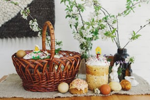 Stylish easter eggs, homemade Easter bread, delicious traditional Easter food in wicker basket and blooming spring flowers on linen napkin on rustic table. Happy Easter! Festive breakfast