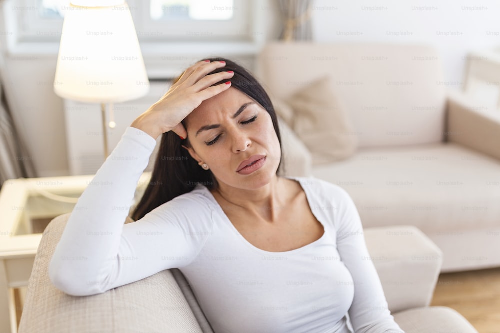 Porträt einer attraktiven Frau, die zu Hause auf einem Sofa sitzt, mit Kopfschmerzen, Schmerzen und einem Ausdruck von Unwohlsein. Verärgerte Frau, die auf der Couch sitzt und starke Kopfschmerzen Migräne verspürt.