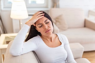 Portrait d’une femme séduisante assise sur un canapé à la maison avec un mal de tête, ressentant de la douleur et avec une expression de malaise. Femme bouleversée assise sur le canapé ressentant un fort mal de tête, migraine.