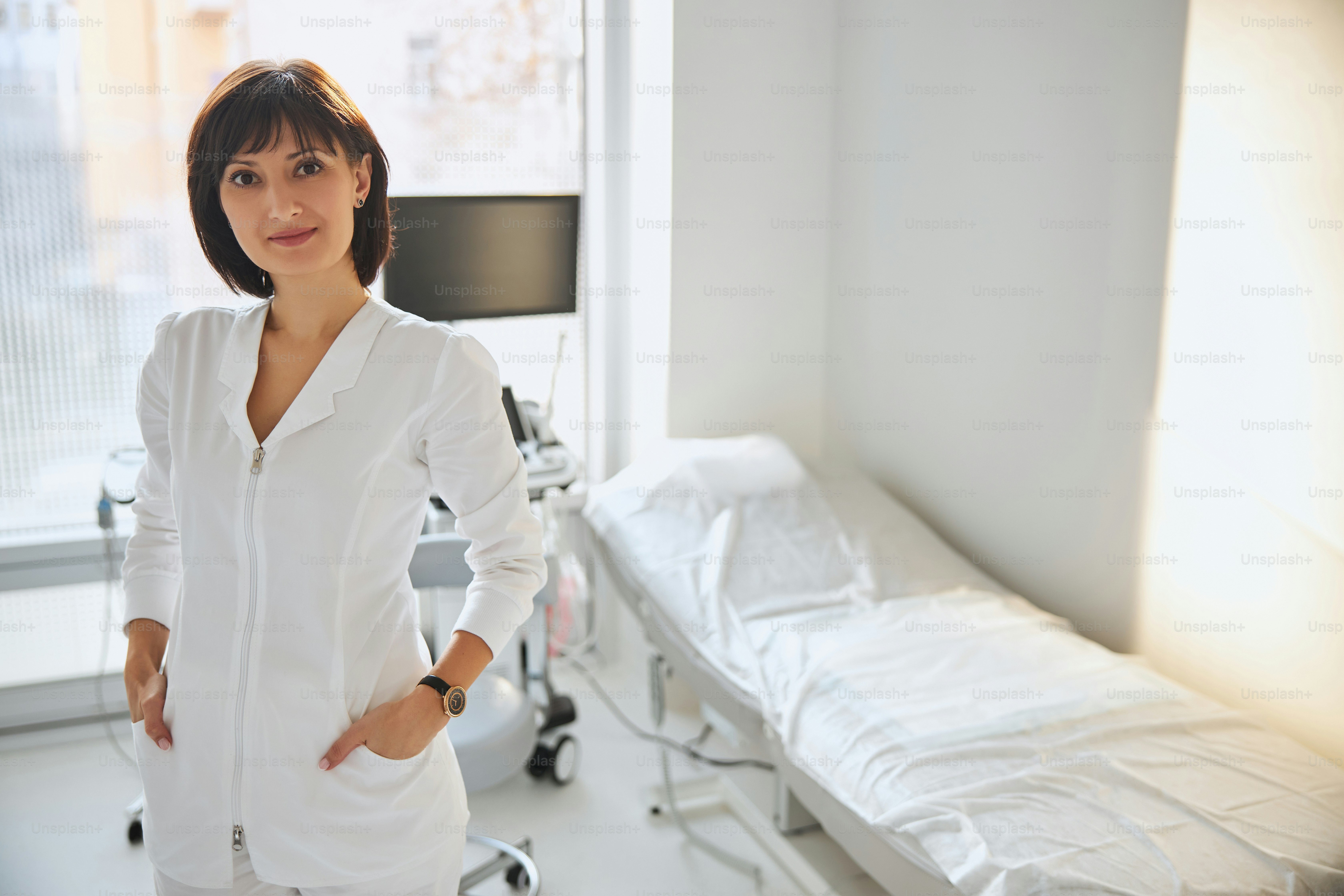 Charmante femme médecin aux cheveux bruns avec ses mains dans les poches de son uniforme debout dans son bureau