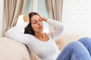Retrato de uma mulher atraente sentada em um sofá em casa com dor de cabeça, sentindo dor e com uma expressão de estar doente. Mulher deprimida chateada deitada no sofá sentindo forte enxaqueca de dor de cabeça.