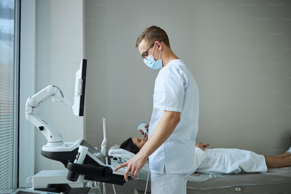 Vista lateral de un médico con gafas con una sonda ultrasónica en una mano mirando la consola del aparato de ecografía