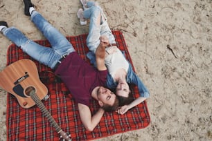 Acoustic guitar near them. Young couple have picnic on the beach. Lying on the red colored blanket.