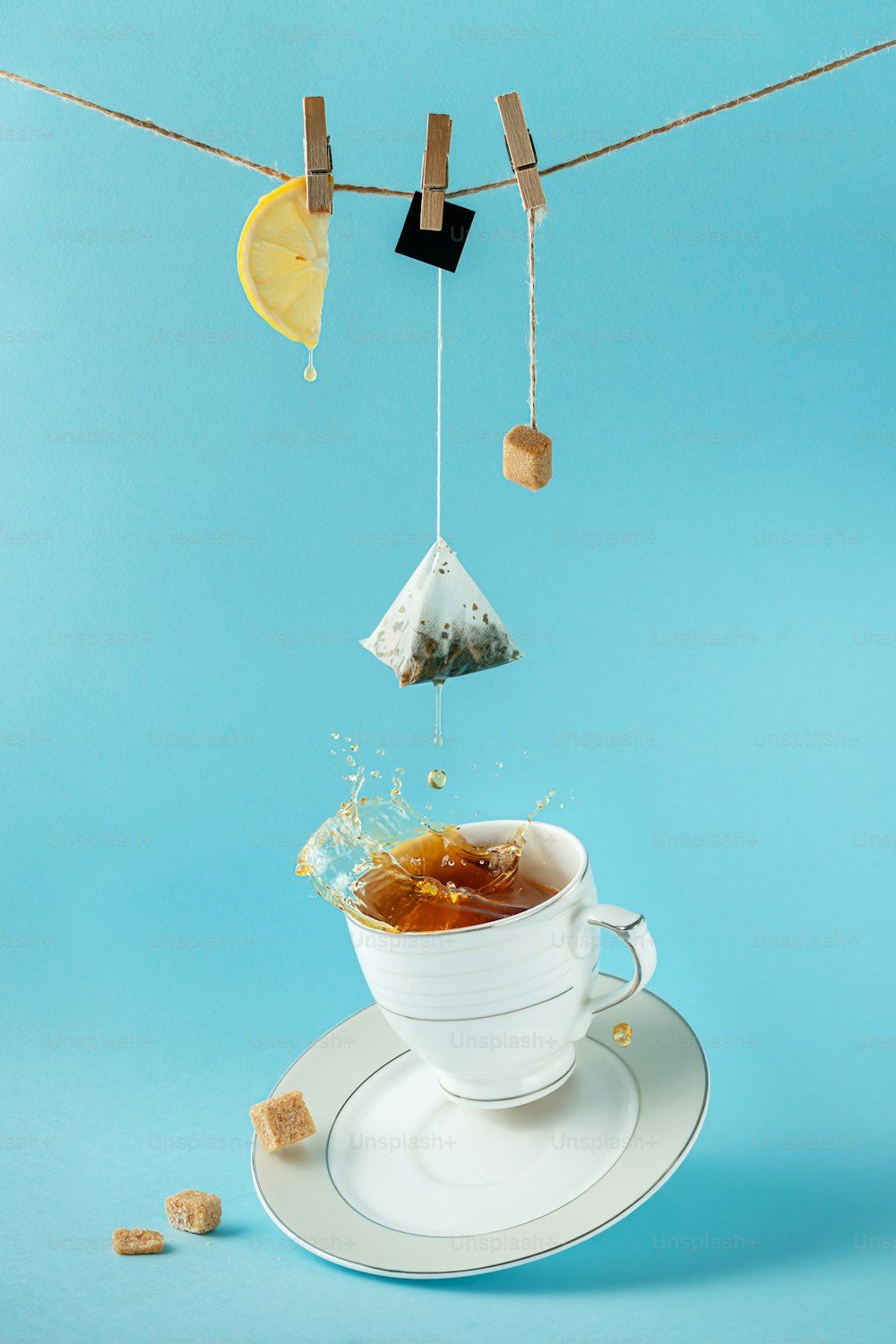 Teebeutel, Zitrone und Zucker hängen am Seil über spritzendem Tee in der Tasse auf blauem Hintergrund. Kreatives Stillleben