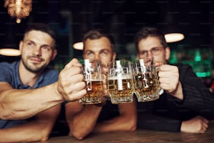 Golpeando vasos. Tres aficionados al deporte en un bar viendo fútbol. Con cerveza en la mano.