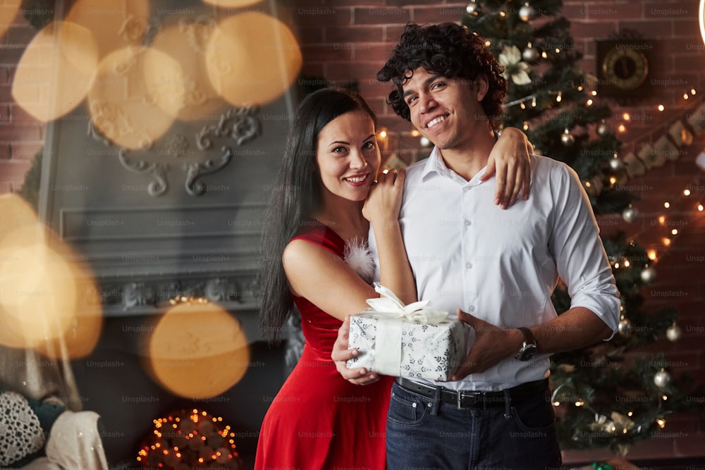 Este regalo es para ti. Hermosa pareja celebrando el año nuevo en la habitación decorada con árbol de Navidad y chimenea detrás.
