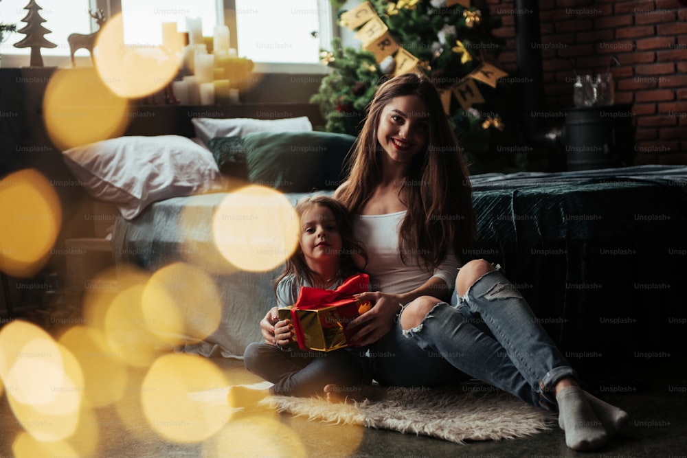 Nice portrait. Mother and daughter sits in holiday decorated room and holds gift box.
