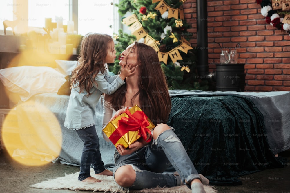 Thankful girl is giving a kiss. Mother and daughter sits in holiday decorated room and holds gift box.