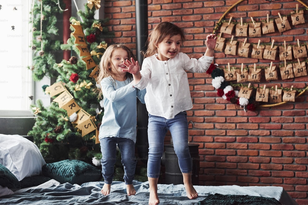 Letters on the tree means merry Christmas. Cheerful kids having fun and jumping on the bed with decorative holiday background.