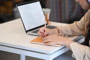 Foto cortada de empresária escrevendo algo em caderno e trabalhando com laptop de computador na mesa do escritório.
