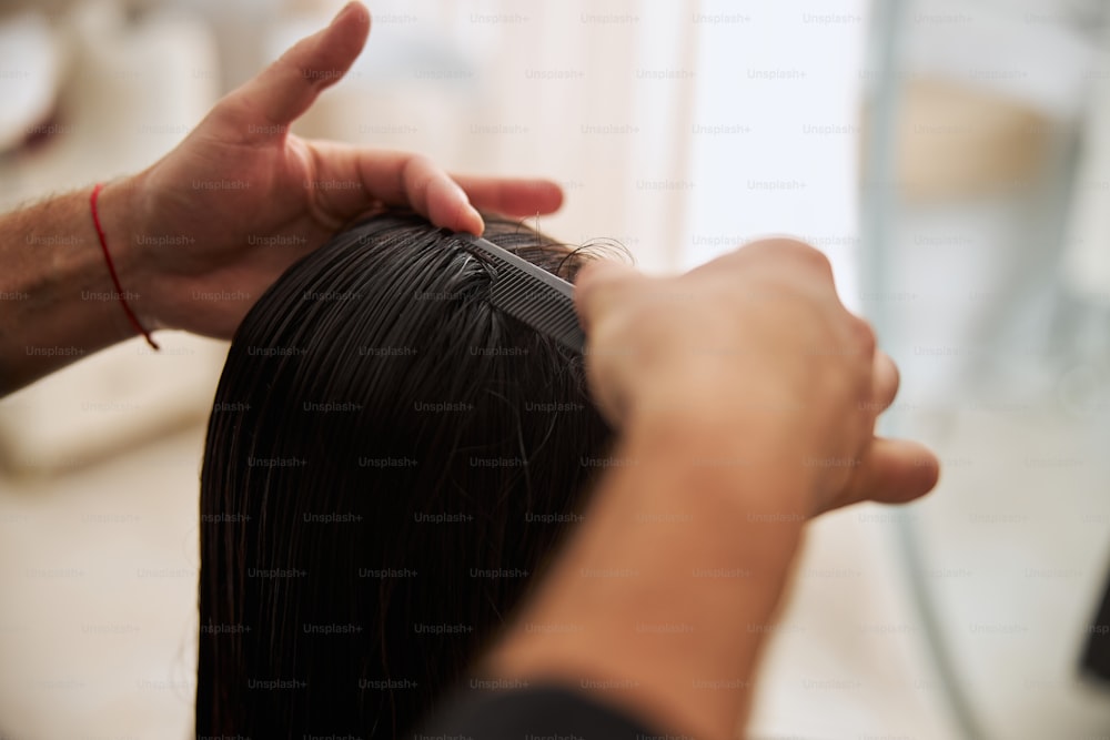 Abgeschnittenes Foto eines Stylisten, der seinen verträumten Kunden mit einem schneidenden Kamm lose dunkle nasse Haare entwirrt