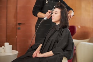 Side view of a calm customer dressed in a hairdressing gown sitting in the barber chair during hairstyling