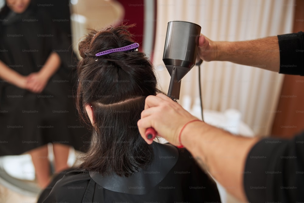 Back view of a dark-haired woman having her hair styled by a coiffeur using a pair of hairdressing tools