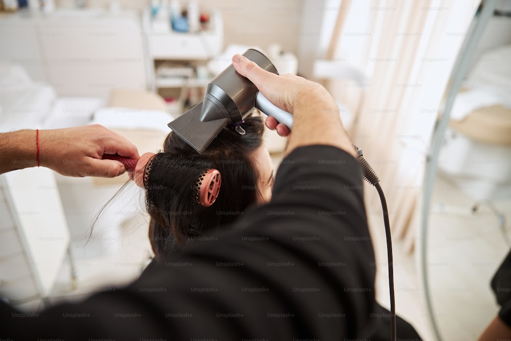 Vista trasera de una clienta con el pelo rizado con un secador de pelo y un cepillo radial