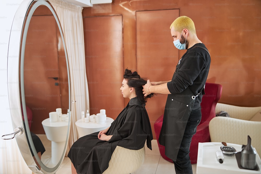 Side view of a straw-haired coiffeur in a face mask sectioning the young woman hair