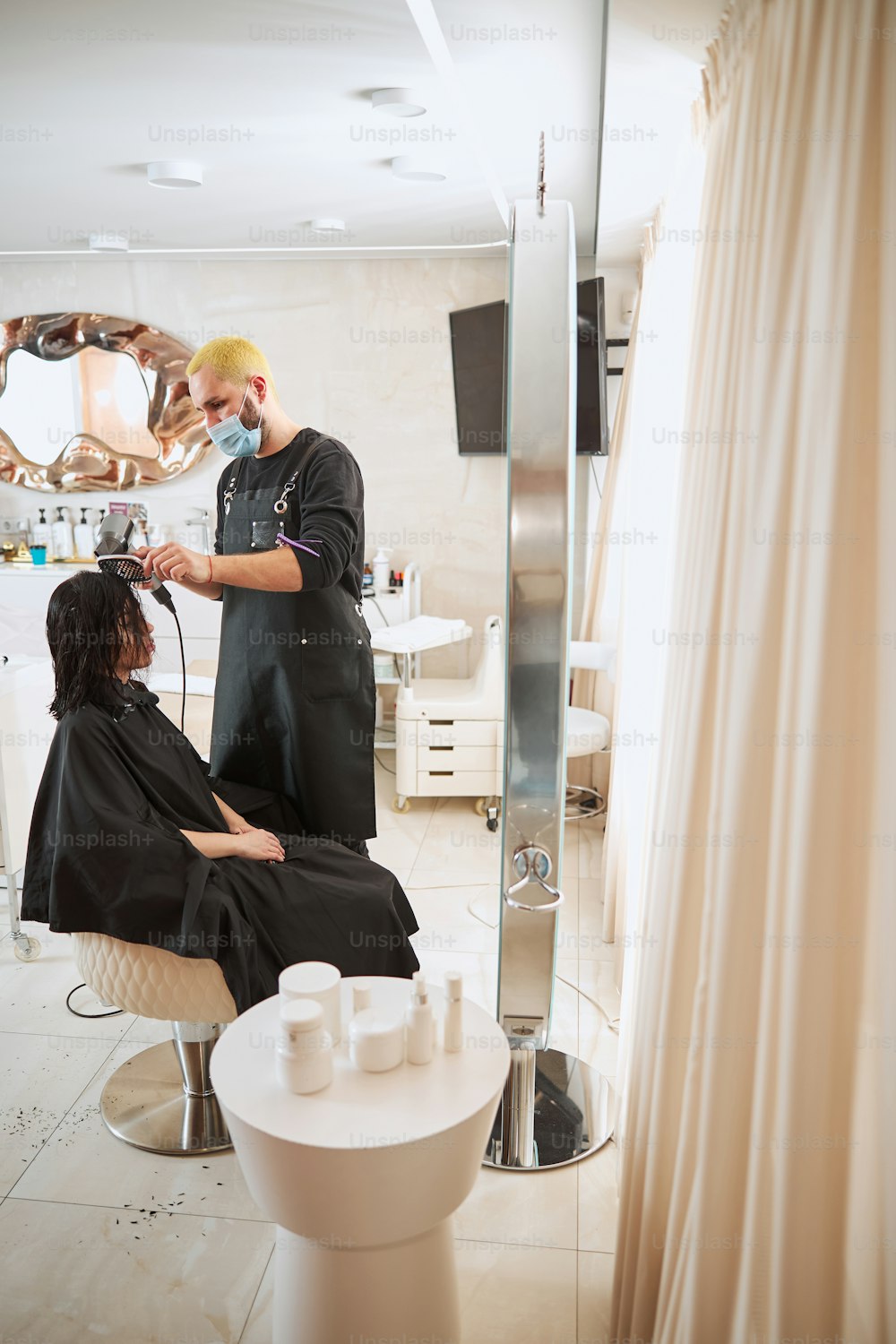 Focused straw-haired hairdresser in a face mask and an apron working on a female hairstyle