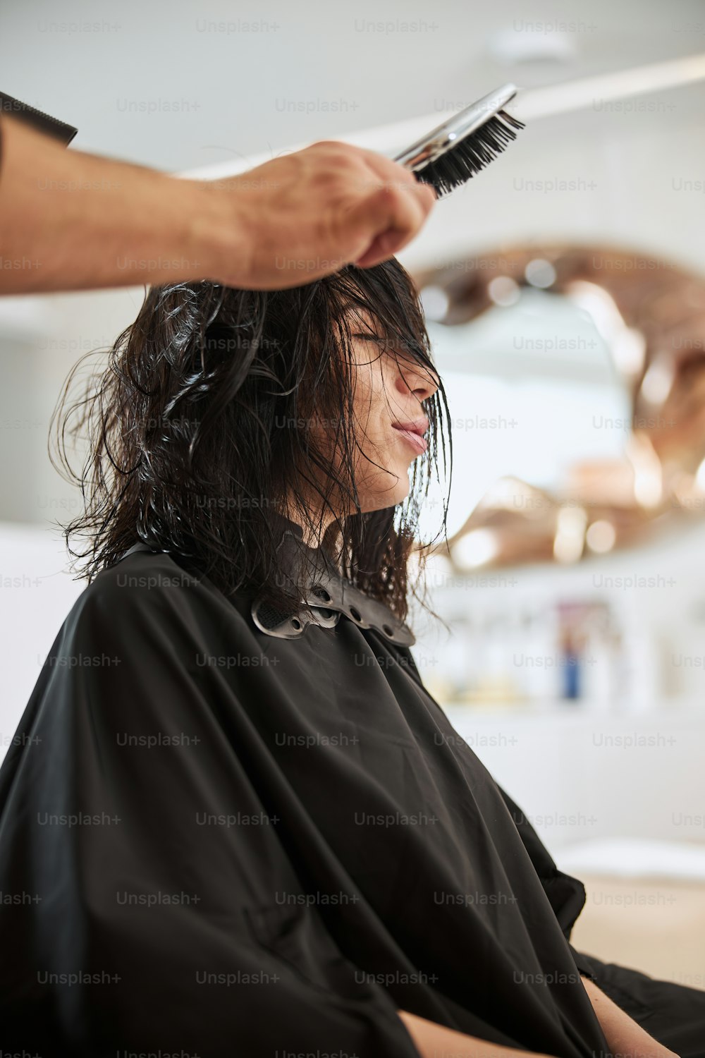 Side view of a serene lady with her eyes closed having her hair styled by a professional hairdresser