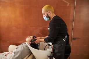 洗髪中に女性の頭皮をマッサージするフェイスマスク姿の美容師の側面図