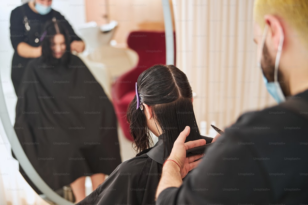Back view of a hairdresser trimming Caucasian lady hair ends using a comb and a pair of scissors