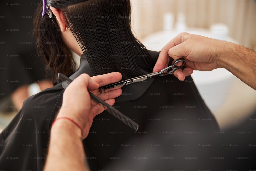Beschnittenes Foto einer dunkelhaarigen Kundin, deren gespaltene Haarspitzen von einem Friseur geschnitten werden