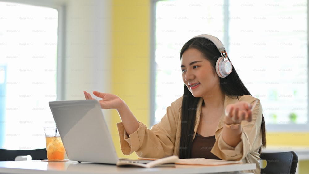 Portrait d’une étudiante avec un casque parlant tout en étudiant en ligne sur un ordinateur portable dans le salon