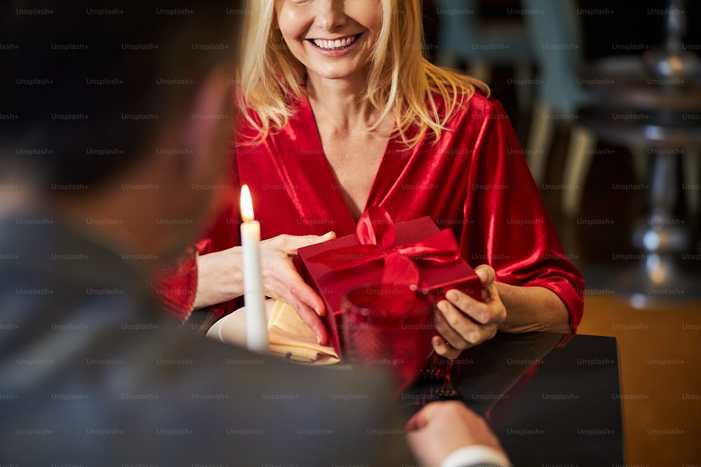 Cropped photo of surprised blonde woman smiling widely while receiving red gift box from man at restaurant table