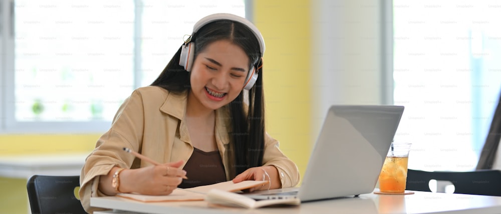 Portrait of young female student with headphone smiling while doing home working with stationery and laptop