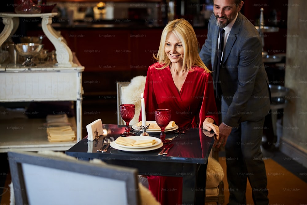 Well-mannered man smiling while helping a blonde woman into her chair at the restaurant
