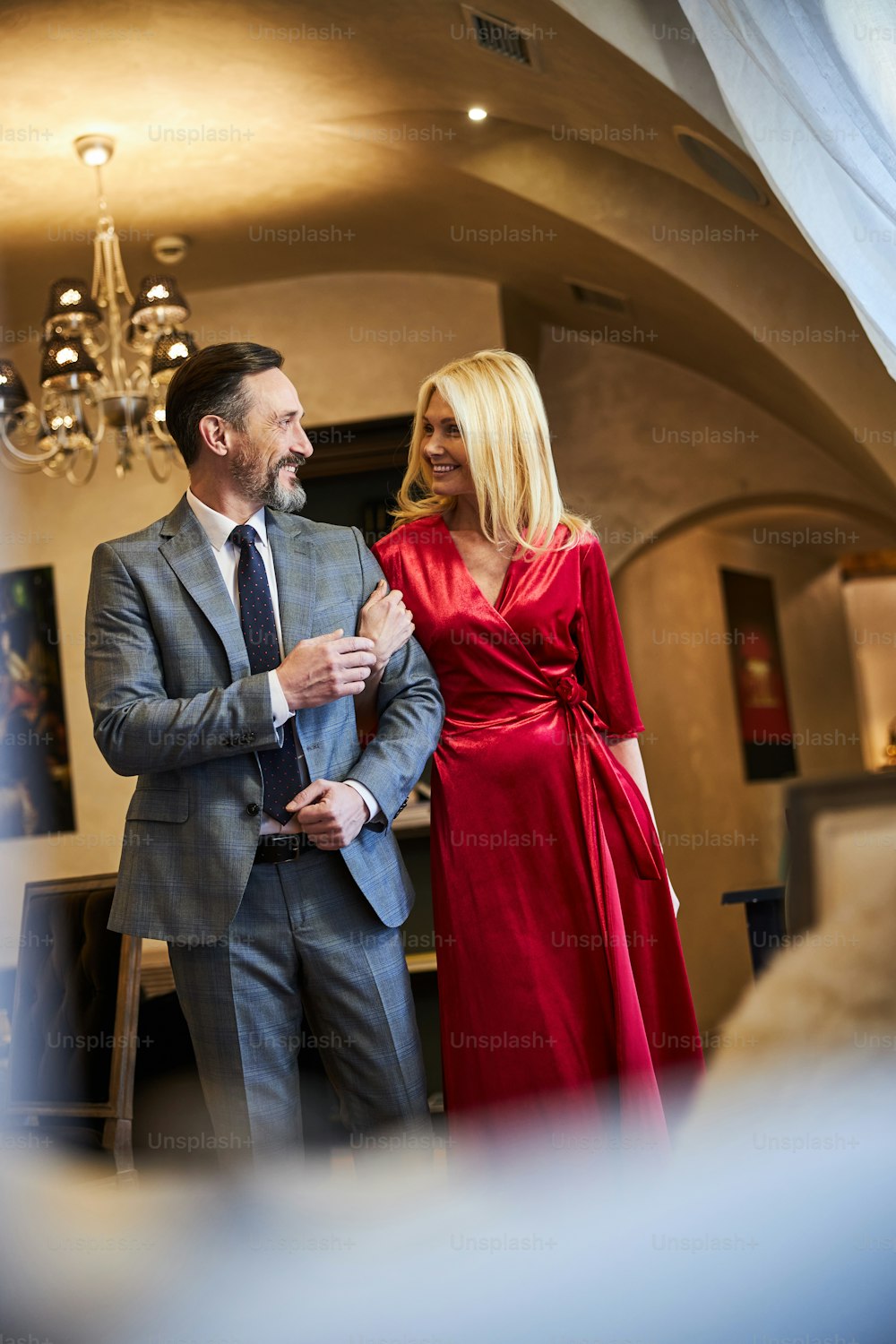 Excited blonde woman in red dress smiling while walking arm in arm with elegant gentleman and looking happy