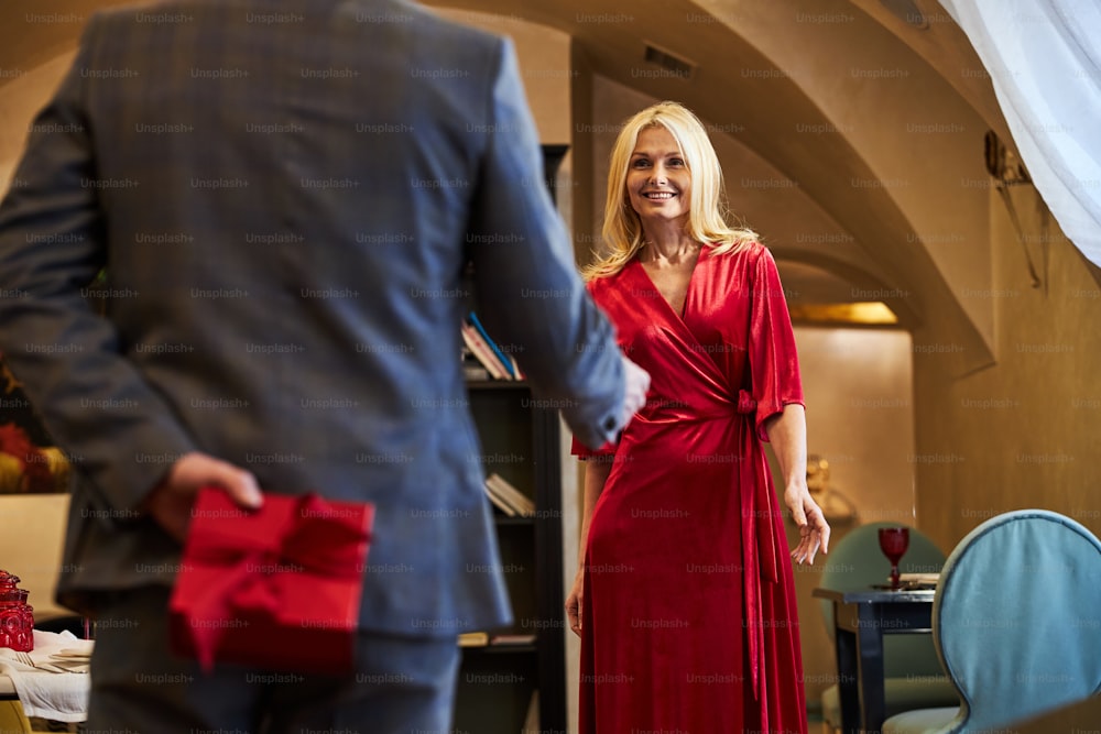 Herr im Anzug hält rote Geschenkbox hinter dem Rücken, um blonde Frau vor sich zu überraschen