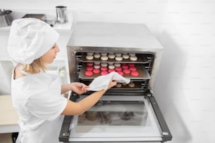Confectionery shop, macarons baking. Pretty woman confectioner in white uniform and hat, holds a batch of macarons on baking tray, ready to take it out of the oven after baking