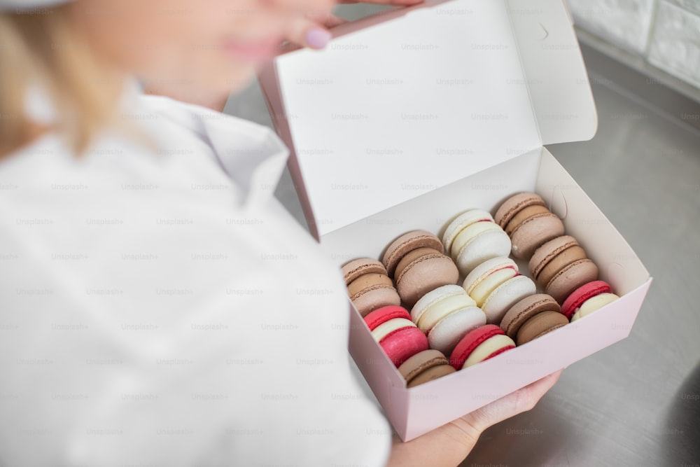 Close up top angle view of colorful macarons in a gift box in hands of female confectioner. Traditional french dessert macaroons in a rows in paper present box.