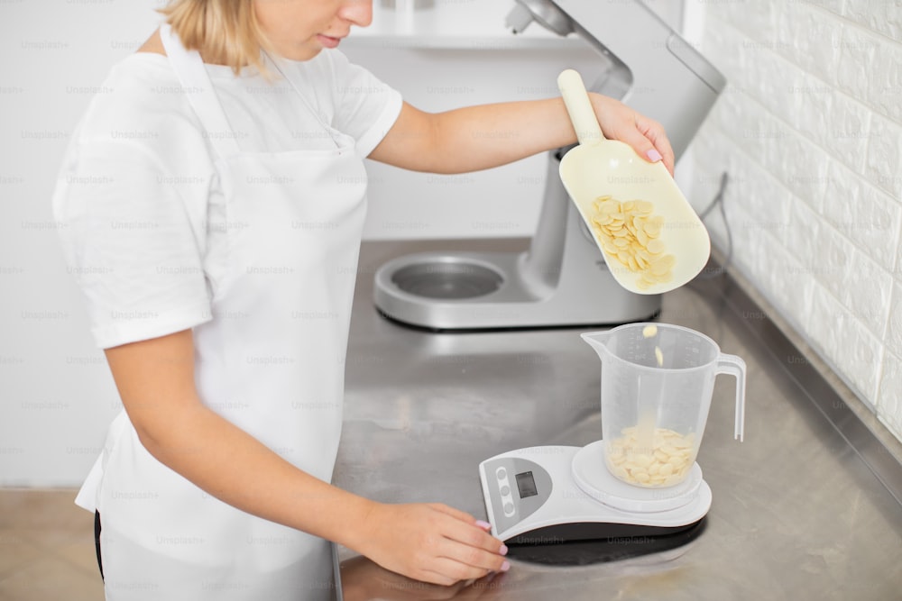Confectionery shop, baking concept. Close up cropped image of young professional female pastry chef, weighing ingredients for ganache or cream, putting white chocolate drops in measuring cup.