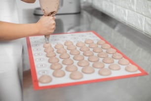 O processo de aplicação de uma massa de macarons em esteira de silicone. Close up foto cortada de mãos femininas segurando saco de pastelaria, espremendo massa de amêndoa marrom em um tapete de silicone de cozimento.