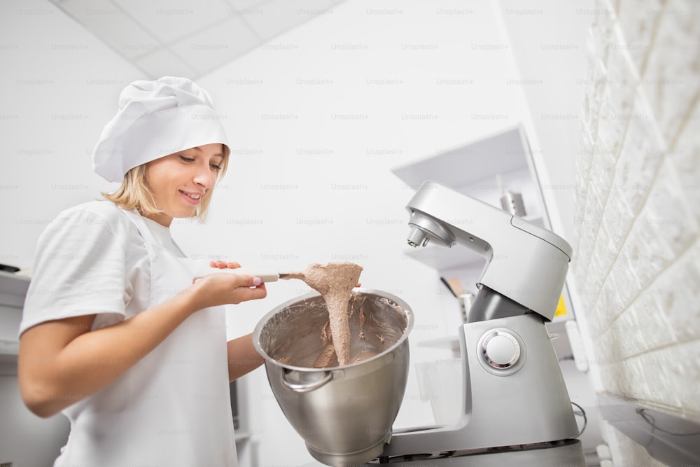 Close up side view photo of young girl confectioner chef, checking the mixed dough, holding a spatula, mixing the dough for macaroons in a metal bowl.