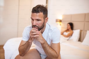 Young couple having a problem. Guy is sitting on bed and looking sadly away, his girlfriend in the background. Upset young couple having problems with sex