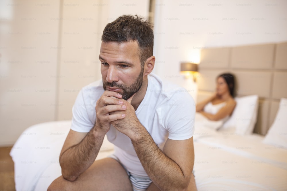 問題を抱えている若いカップル。男はベッドに座って悲しそうに目をそらし、背後にはガールフレンドがいる。セックスで問題を抱えている動揺した若いカップル