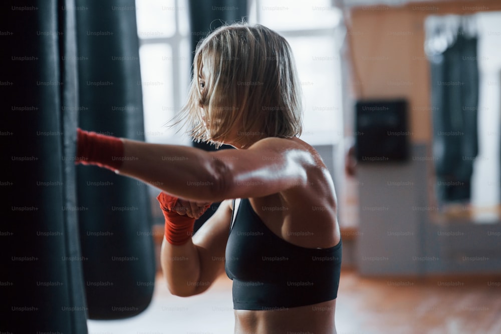 Hair covering the face in moment of kick. Female boxer is punching the bag. Blonde have exercise in the gym.