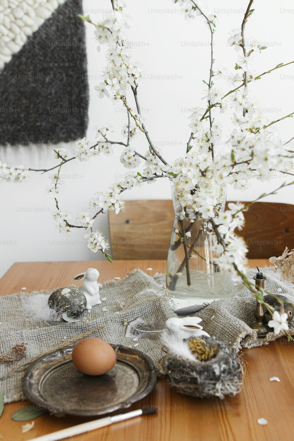 Frohe Ostern! Natürliche Ostereier, weiße Hasen, Federn, Nest und kirschblühender Zweig mit Blütenblättern auf rustikaler Leinenserviette auf Tisch. Platz für Text. Umweltfreundliche ländliche Einrichtung