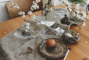 Uova di Pasqua naturali, coniglietti bianchi, piume, nido e ramo fiorito di ciliegio con petali su tovagliolo di lino rustico sul tavolo. Buona Pasqua. Spazio per il testo. Arredamento ecologico