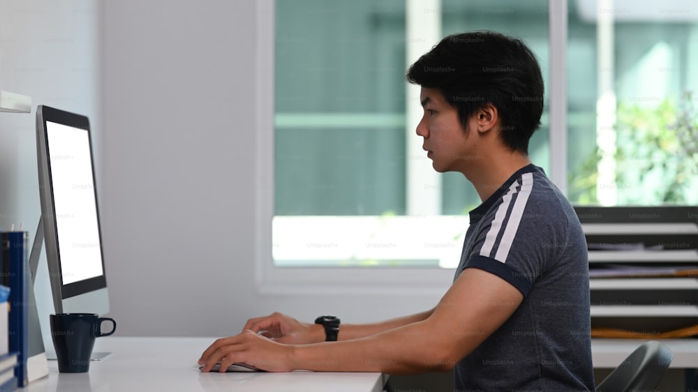 ホームオフィスに座りながら、コンピュータでオンラインで作業している若い男性の側面図。