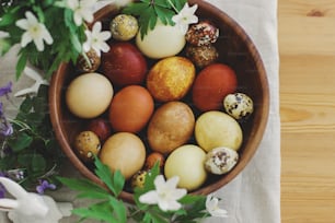 Stylish easter eggs in wooden bowl on rustic table with bunny figurines and spring blooming flowers. Top view with space for text. Happy Easter! Natural dyed eggs in yellow and red colors.