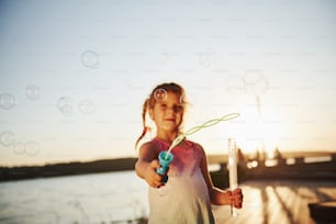 Evening time. Happy little girl playing with bubbles near the lake at park.