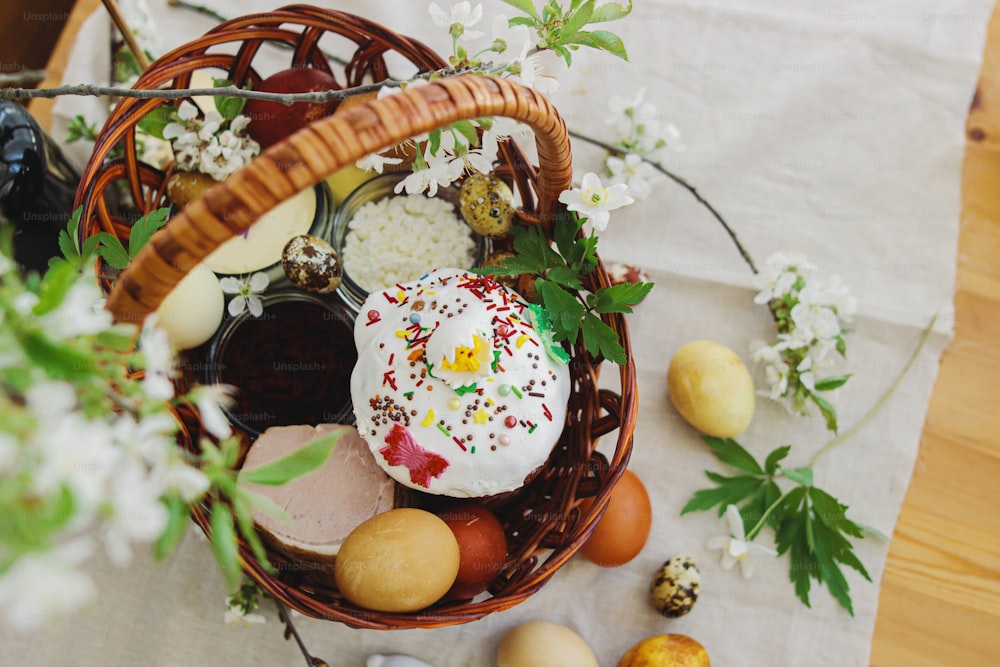 Delicious Easter food, stylish easter eggs, beets, cheese, butter, ham, homemade Easter bread in wicker basket with blooming spring flowers on rustic table, orthodox traditions. Happy Easter!
