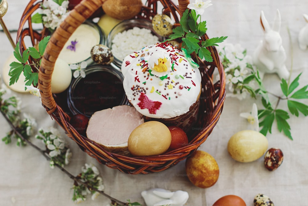 Stylish easter eggs, homemade Easter bread, delicious traditional Easter food in wicker basket and blooming spring flowers on linen napkin on rustic table. Happy Easter! Festive breakfast