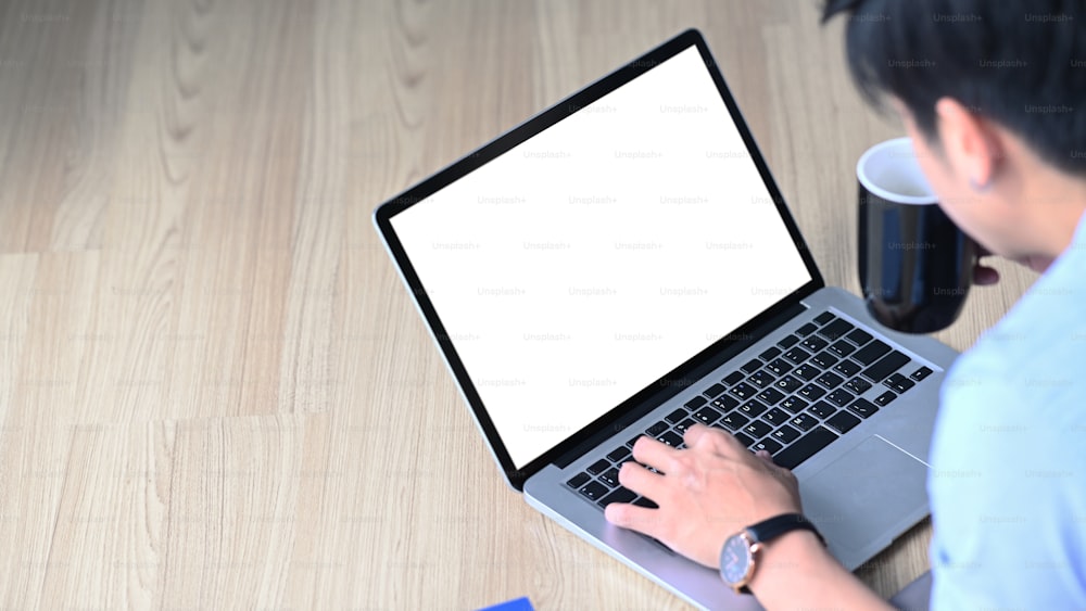 Close up view of young man drinking coffee and using computer laptop while lying on wooden floor.