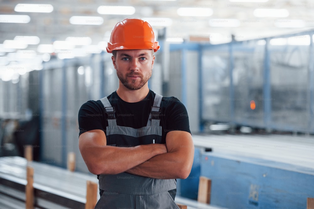 Fica de braços cruzados. Retrato do trabalhador industrial do sexo masculino dentro de casa na fábrica. Jovem técnico com capacete laranja.