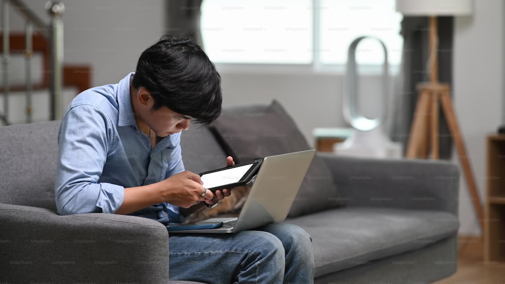 Young asian man freelancer working on laptop and using digital tablet and while sitting on couch at home.